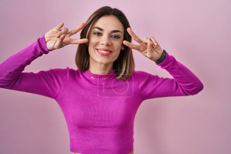 Téléchargez les photos : Hispanic woman standing over pink background doing peace symbol with fingers over face, smiling cheerful showing victory - en image libre de droit