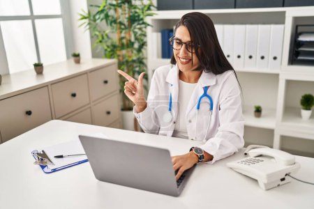 Photo for Young hispanic doctor woman doing video call at the clinic smiling happy pointing with hand and finger to the side - Royalty Free Image