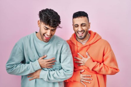 Foto de Young hispanic gay couple standing over pink background smiling and laughing hard out loud because funny crazy joke with hands on body. - Imagen libre de derechos