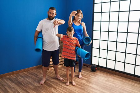 Photo for Family of three holding yoga mat with angry face, negative sign showing dislike with thumbs down, rejection concept - Royalty Free Image
