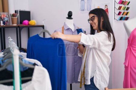 Photo for Young beautiful hispanic woman tailor make photo by smartphone to sweater at atelier - Royalty Free Image