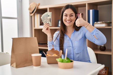 Photo for Young brunette woman eating take away food at home holding money smiling happy and positive, thumb up doing excellent and approval sign - Royalty Free Image
