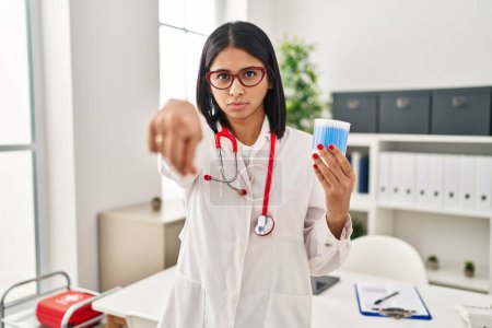 Foto de Young hispanic doctor woman holding cotton buds pointing with finger to the camera and to you, confident gesture looking serious - Imagen libre de derechos
