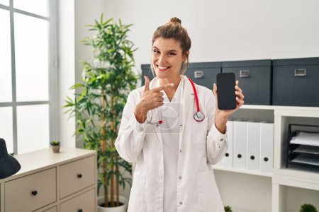 Téléchargez les photos : Young woman wearing doctor uniform holding smartphone smiling happy pointing with hand and finger - en image libre de droit