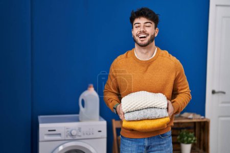 Téléchargez les photos : Hispanic man with beard holding clean folded laundry smiling and laughing hard out loud because funny crazy joke. - en image libre de droit