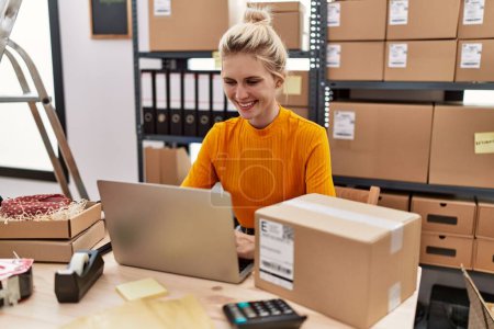 Téléchargez les photos : Young blonde woman working at small business ecommerce using laptop looking positive and happy standing and smiling with a confident smile showing teeth - en image libre de droit