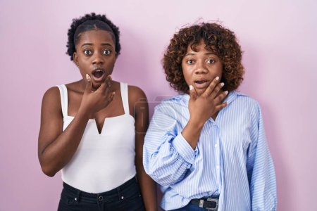 Photo for Two african women standing over pink background looking fascinated with disbelief, surprise and amazed expression with hands on chin - Royalty Free Image