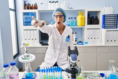 Photo for Brunette woman working at scientist laboratory pointing with finger surprised ahead, open mouth amazed expression, something on the front - Royalty Free Image