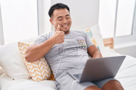 Photo for Chinese young man using laptop sitting on the bed smiling happy and positive, thumb up doing excellent and approval sign - Royalty Free Image