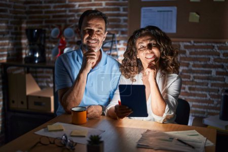 Photo for Middle age hispanic couple using touchpad sitting on the table at night with hand on chin thinking about question, pensive expression. smiling and thoughtful face. doubt concept. - Royalty Free Image