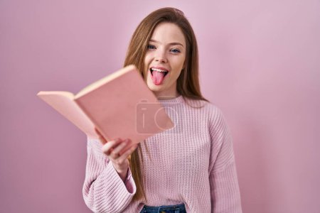 Foto de Joven mujer caucásica leyendo un libro sobre fondo rosa sacando la lengua feliz con expresión divertida. - Imagen libre de derechos