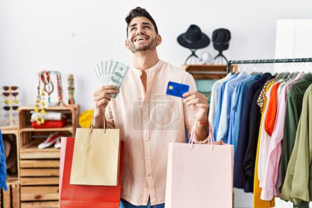 Foto de Joven hispano sosteniendo bolsas de compras y tarjeta de crédito sonriendo y riendo en voz alta porque divertido chiste loco. - Imagen libre de derechos
