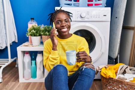 Foto de Mujer afroamericana hablando en el teléfono inteligente bebiendo café esperando la lavadora en la sala de lavandería - Imagen libre de derechos