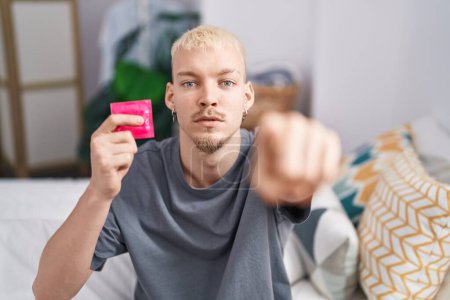 Foto de Young caucasian man holding condom sitting on bed pointing with finger to the camera and to you, confident gesture looking serious - Imagen libre de derechos