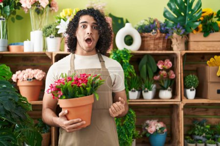 Téléchargez les photos : Homme hispanique avec les cheveux bouclés travaillant à la boutique de fleuriste tenant l'usine effrayée et étonnée avec la bouche ouverte pour la surprise, visage incrédule - en image libre de droit