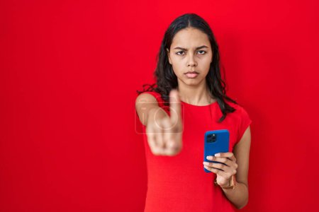 Téléchargez les photos : Jeune femme brésilienne à l'aide d'un smartphone sur fond rouge pointant du doigt et l'expression en colère, ne montrant aucun geste - en image libre de droit