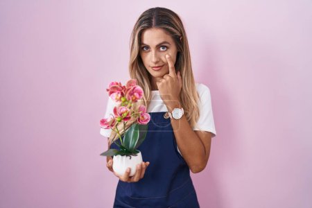 Foto de Joven mujer rubia vistiendo delantal jardinero sosteniendo planta apuntando al ojo observándote gesto, expresión sospechosa - Imagen libre de derechos