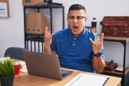 Téléchargez les photos : Jeune homme hispanique travaillant au bureau avec un ordinateur portable criant avec une expression folle faisant symbole rock avec les mains en l'air. star de la musique. concept lourd. - en image libre de droit