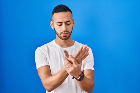 Téléchargez les photos : Jeune homme hispanique debout sur fond bleu souffrant de douleur aux mains et aux doigts, inflammation de l'arthrite - en image libre de droit