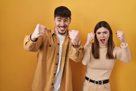 Foto de Young hispanic couple standing over yellow background angry and mad raising fists frustrated and furious while shouting with anger. rage and aggressive concept. - Imagen libre de derechos
