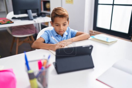 Téléchargez les photos : Adorable hispanic boy student sitting on table doing homework at classroom - en image libre de droit