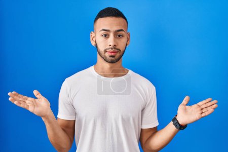 Foto de Young hispanic man standing over blue background clueless and confused expression with arms and hands raised. doubt concept. - Imagen libre de derechos