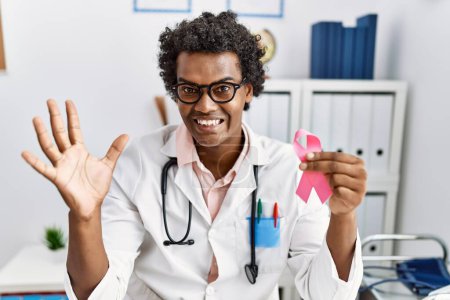 Photo for African doctor man holding pink cancer ribbon celebrating victory with happy smile and winner expression with raised hands - Royalty Free Image