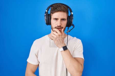 Téléchargez les photos : Homme hispanique avec barbe écoutant de la musique portant un casque d'écoute fasciné par l'incrédulité, la surprise et l'expression étonnante avec les mains sur le menton - en image libre de droit