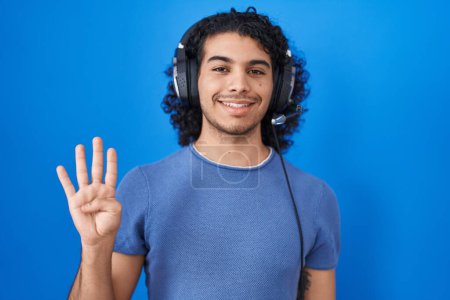 Téléchargez les photos : Hispanic man with curly hair listening to music using headphones showing and pointing up with fingers number four while smiling confident and happy. - en image libre de droit