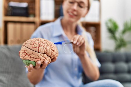 Téléchargez les photos : Jeune femme blonde psychologue tenant le cerveau assis sur le canapé à la clinique - en image libre de droit
