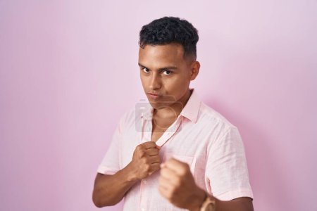 Photo for Young hispanic man standing over pink background ready to fight with fist defense gesture, angry and upset face, afraid of problem - Royalty Free Image