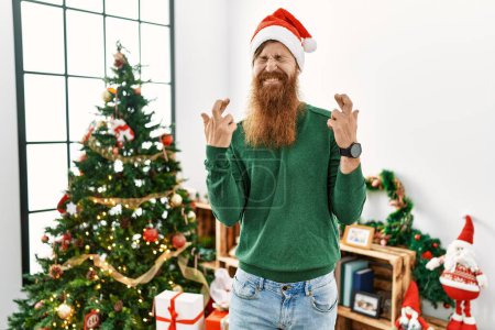 Photo for Redhead man with long beard wearing christmas hat by christmas tree gesturing finger crossed smiling with hope and eyes closed. luck and superstitious concept. - Royalty Free Image