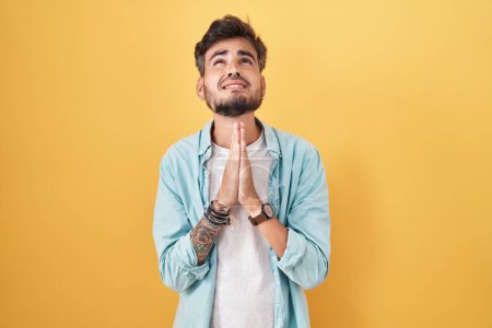 Téléchargez les photos : Young hispanic man with tattoos standing over yellow background begging and praying with hands together with hope expression on face very emotional and worried. begging. - en image libre de droit