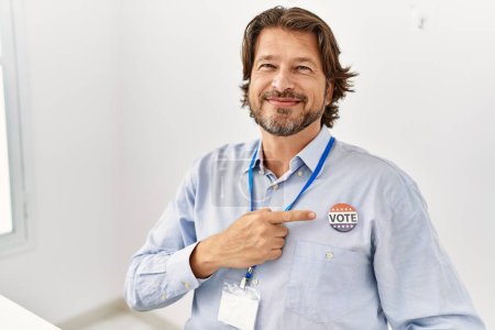 Foto de Middle age caucasian man smiling confident pointing with finger to badge at electoral college - Imagen libre de derechos