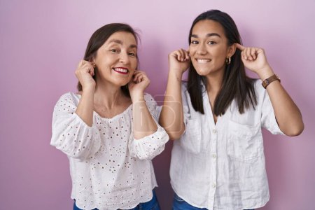 Foto de Madre e hija hispanas juntas sonriendo tirando de las orejas con los dedos, gesto divertido. problema de audición - Imagen libre de derechos
