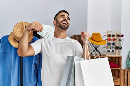Foto de Young hispanic man customer holding shopping bags and faning with dollars at clothing store - Imagen libre de derechos