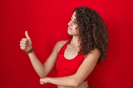 Téléchargez les photos : Hispanic woman with curly hair standing over red background looking proud, smiling doing thumbs up gesture to the side - en image libre de droit