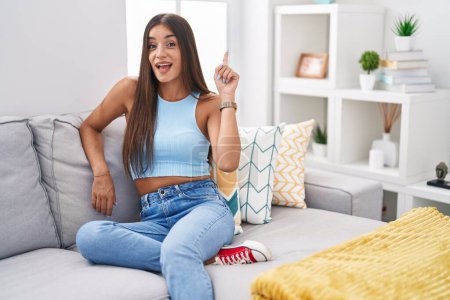 Téléchargez les photos : Jeune femme brune assise sur le canapé à la maison pointant du doigt avec une idée réussie. Sorti et heureux. numéro un. - en image libre de droit