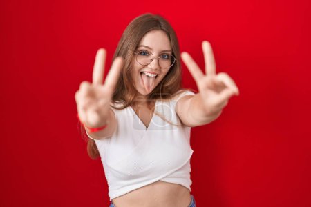 Téléchargez les photos : Jeune femme caucasienne debout sur fond rouge souriant avec la langue montrant les doigts des deux mains faisant signe de victoire. numéro deux. - en image libre de droit