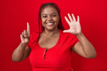 Foto de Mujer afroamericana con el pelo trenzado de pie sobre fondo rojo mostrando y apuntando hacia arriba con los dedos número seis mientras sonríe confiado y feliz. - Imagen libre de derechos