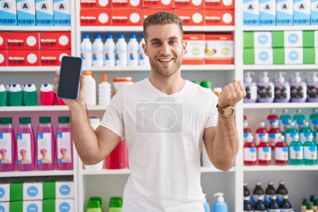 Photo for Young caucasian man working at pharmacy drugstore showing smartphone screen screaming proud, celebrating victory and success very excited with raised arm - Royalty Free Image