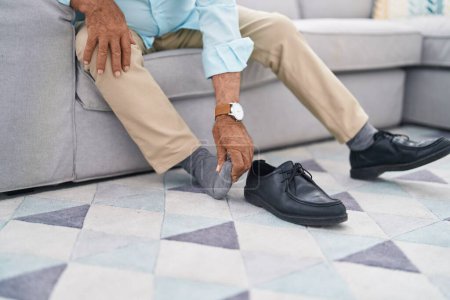 Foto de Senior grey-haired man suffering for feet pain sitting on sofa at home - Imagen libre de derechos