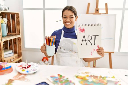 Photo for Young latin woman holding art notebook and cup with pencils at art studio - Royalty Free Image