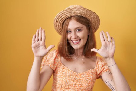 Foto de Joven pelirroja de pie sobre fondo amarillo con sombrero de verano mostrando y señalando hacia arriba con los dedos número diez mientras sonríe confiado y feliz. - Imagen libre de derechos