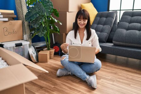 Foto de Young brunette woman sitting on the floor with laptop at new home smiling happy pointing with hand and finger - Imagen libre de derechos