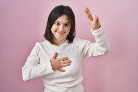 Foto de Woman with down syndrome standing over pink background smiling swearing with hand on chest and fingers up, making a loyalty promise oath - Imagen libre de derechos