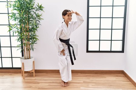 Photo for Young hispanic girl wearing karate kimono and black belt very happy and smiling looking far away with hand over head. searching concept. - Royalty Free Image