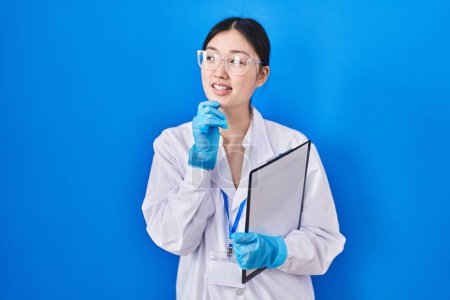 Foto de Chinese young woman working at scientist laboratory with hand on chin thinking about question, pensive expression. smiling and thoughtful face. doubt concept. - Imagen libre de derechos