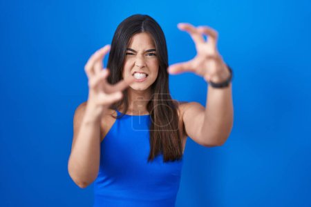 Foto de Mujer hispana de pie sobre fondo azul gritando frustrada por la rabia, manos tratando de estrangular, gritando loco - Imagen libre de derechos