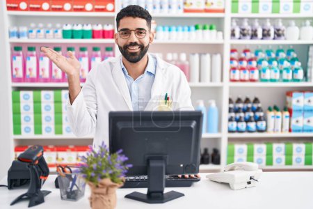 Téléchargez les photos : Hispanic man with beard working at pharmacy drugstore smiling cheerful presenting and pointing with palm of hand looking at the camera. - en image libre de droit